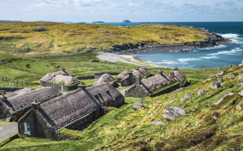 Aerial view of Hebrides, Scotland, with rugged coast, grassy hills & dark brown barn-like buildings nestled together.