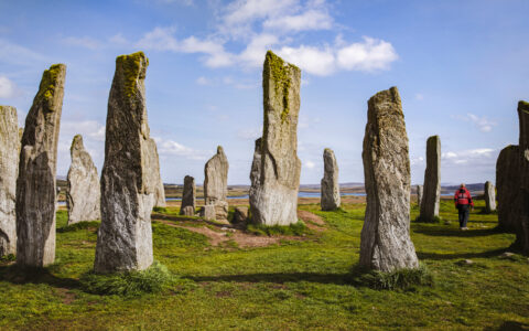 Scottish Isles & Norwegian Fjords cruise guest in red coat walks green grass among a monolith of standing stones under blue sky.