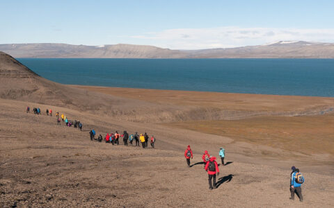Line of South Spitsbergen cruise guests in various colorful coats hike over arid tundra near the sea on a sunny day.