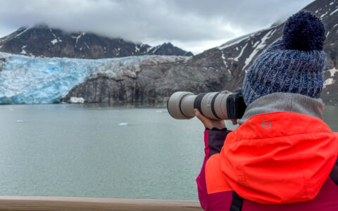 Svalbard Express cruise guest in red jacket & blue knit hat holds zoom lens over railing to photograph an icy blue glacier.