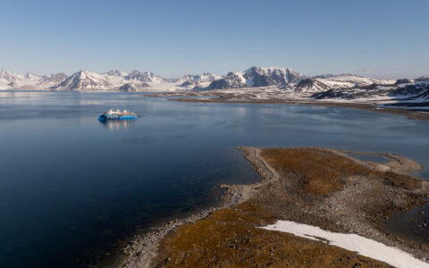 Modern small ship painted blue sits in glassy sea beside small spit of land backed by sharp snowy peaks in Svalbard.