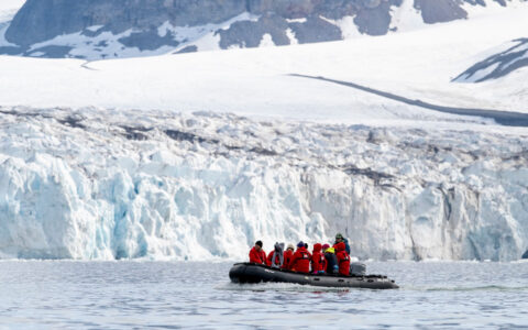 Zodiac boat with Svalbard travelers in red outerwear moves through calm water beside large white & blue glacier in the sun.