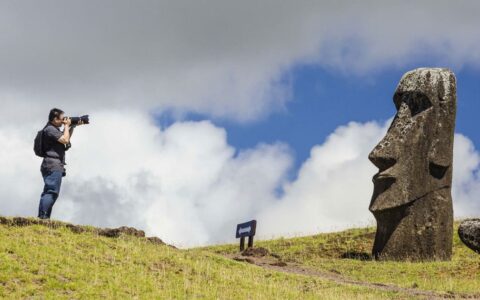solo traveler takes a photograph of a rapa nui statue in easter island chile while standing on a grassy knoll on a partly sunny day