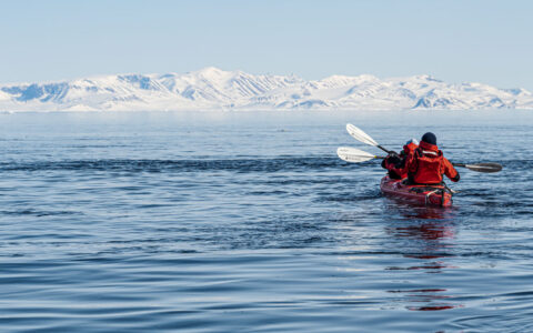 Tandem kayakers in red boat & parkas paddle calm sea beside snow-covered landscapes on a sunny day in Greenland.