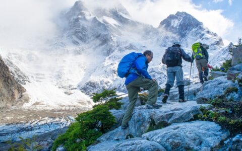 3 trekkers hike up white boulders on a sunny day with massive peaks in the background during the Torres del Paine Trek.