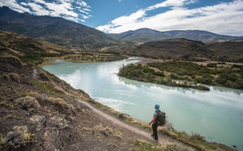 Man stands on smooth dirt trail overlooking a milky turquoise glacial lake on a sunny day during the Torres Del Paine Trek.