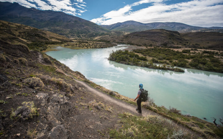 Man stands on smooth dirt trail overlooking a milky turquoise glacial lake on a sunny day during the Torres Del Paine Trek.