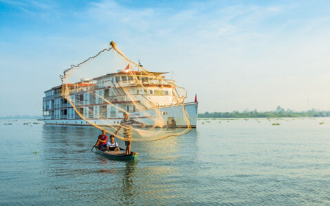 Jahan southeast asia cruise ship with white decks & red trim sits behind canoe of 3 fishermen casting a net on a sunny day,