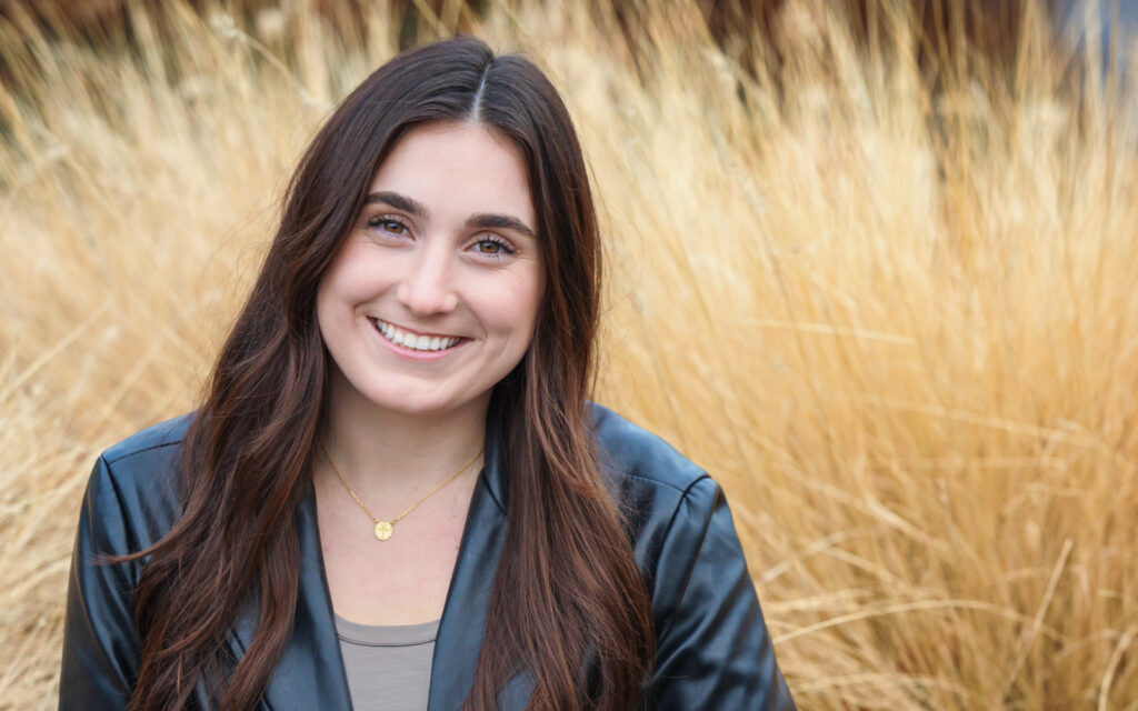 Portrait of Grace Bosco showing her wearing a black blazer, brown top and gold necklace. She's posing in front of a brown plant.