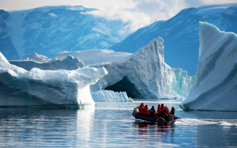 A zodiac navigates large icebergs in calm still waters with snowy mountains off in the distance