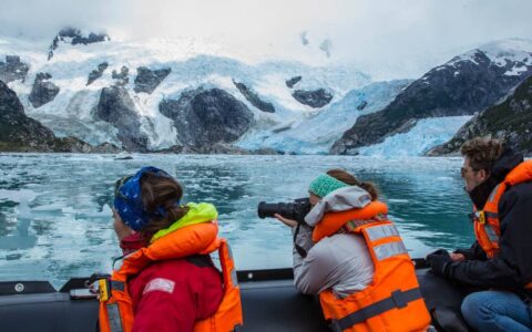 adventure travelers sitting on the edge of a small ship taking pictures across the water of patagonia fjords