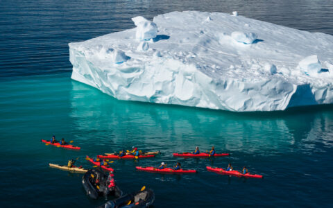 Kayakers in red and yellow kayaks stopped next to a large iceberg floating in the middle of the ocean