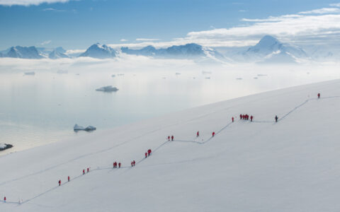 Travelers hike up a snowy hill in a zig zag position in front of breathtaking mountains surrounded by fog