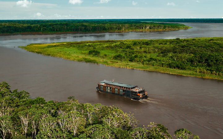 The Aqua Nera cruise ship seen from an aerial shot among a winding Amazon River with bright green trees and bushes along the riverbank