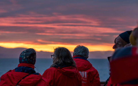 Travelers in red jackets watch the pink and yellow sunset off small cruise ship
