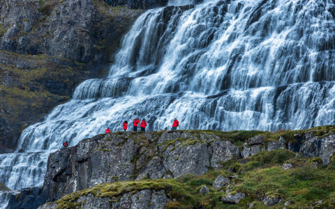 Hikers dressed in red jackets navigate massive water walls that tower over them and fill the whole wall in white water