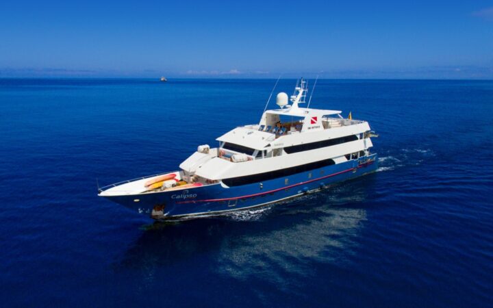 Aerial view of Calipso yacht in Galapagos with dark blue hull & white upper decks, cruising through calm blue water on a sunny day.