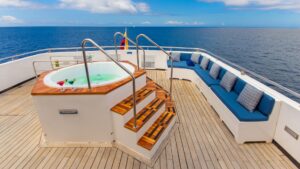 Round Jacuzzi in middle of teak Sundeck with 3 steps & metal handrails beside long blue padded couch aboard the Calipso yacht.