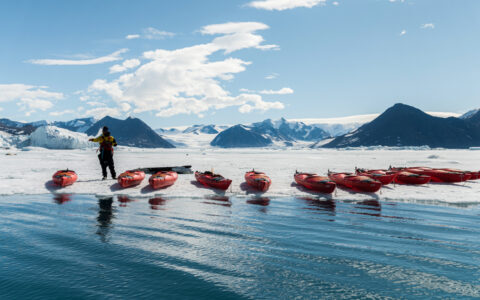 Small fleet of empty red kayaks lined up at water & ice's edge in the sun on the Commandant Charcot Unexplored Antarctica cruise.