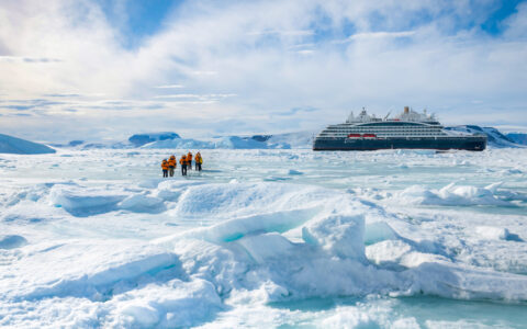 Small group of Unexplored Antarctica cruise guests in orange parkas walk over bumpy ice field near a small ship on a sunny day.