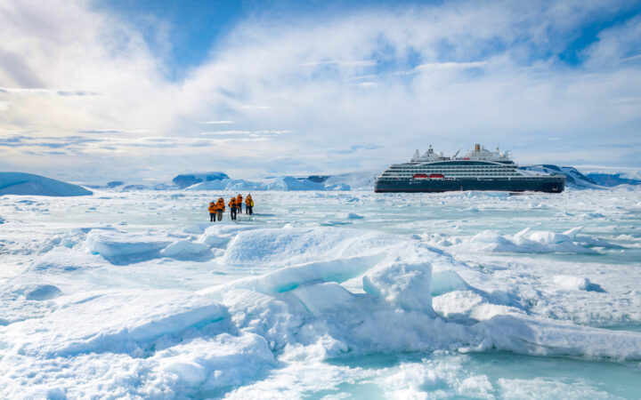 Small group of Unexplored Antarctica cruise guests in orange parkas walk over bumpy ice field near a small ship on a sunny day.