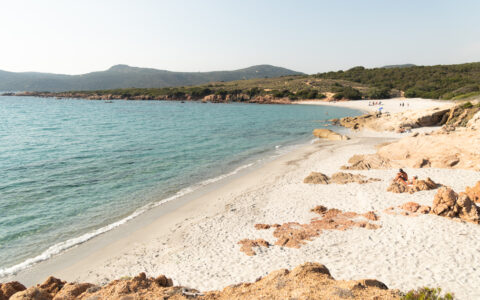 Corsica cruise travelers lay on white-sand beach with beige rocks beside calm teal sea near green hills in the sun.
