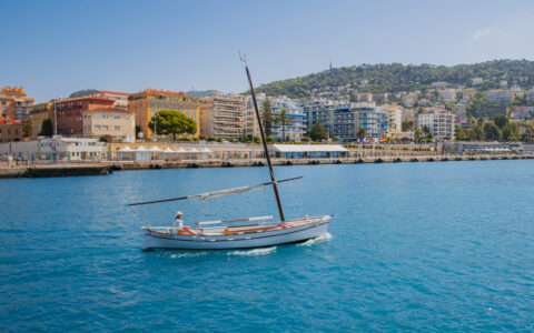 Woman in small white sailboat motors through calm bay with colorful buildings on a sunny day in Nice, France.