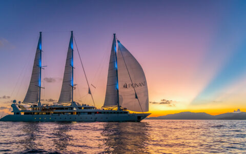 Le Ponant white motorsailor with 3 sails at full mast cruises in calm sea under a purple, blue, orange & yellow sunset.