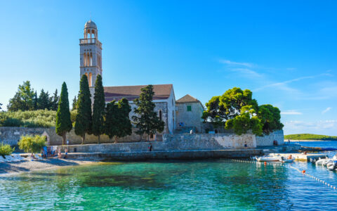 Turquoise waters by small beach & old stone building with clock tower & sea wall, seen on the Croatia Under Sail cruise.