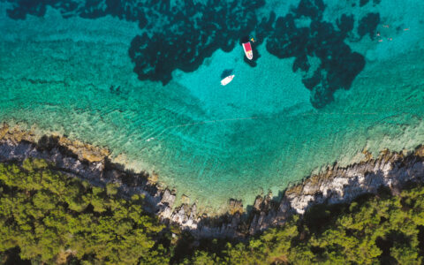 Aerial view looking down onto turquoise waters with Zodiac & swimmers beside green forested shoreline with white rocks in Croatia.