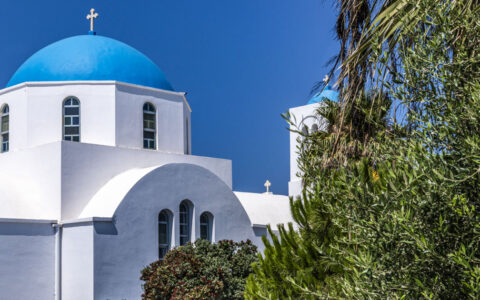 Whitewashed Greek church with blue dome & silver cross on top sits by green foliage in the sun on a Le Ponant Cyclades cruise.