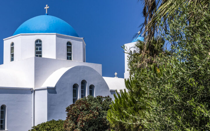 Whitewashed Greek church with blue dome & silver cross on top sits by green foliage in the sun on a Le Ponant Cyclades cruise.