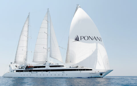 Medium-sized white sailboat Le Ponant with 4 large white sails full of wind sails through calm blue sea in the sun in Greece.