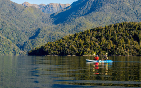 Tandem kayakers paddle a blue boat in calm sea by verdant green hills & peaks in the sun on a Doubtful Sound overnight cruise.