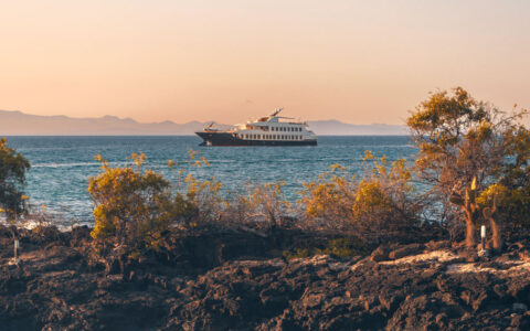 Evolve Galapagos yacht with dark blue hull & 2 white upper decks sits in calm water by rocky small island at sunset.