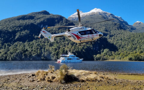 Red & white helicopter flies above small white yacht on a sunny day among dense green hills during a fiordland cruise.