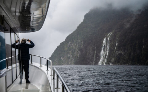 Man stands on deck of small white yacht & photographs large waterfall coming down from green cliffs in New Zealand.