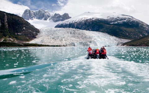 Zodiac boat with Patagonia cruise guests in orange life jackets motors toward Pia Glacier with jagged peaks behind in the sun.