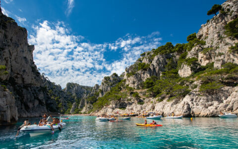 French Riviera cruise guests swim & kayak from small craft in teal sea among beige cliffs with green pine forest in the sun.