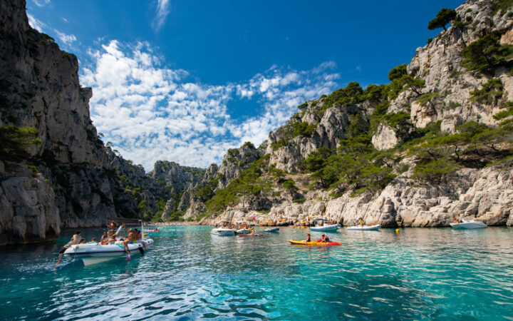 French Riviera cruise guests swim & kayak from small craft in teal sea among beige cliffs with green pine forest in the sun.