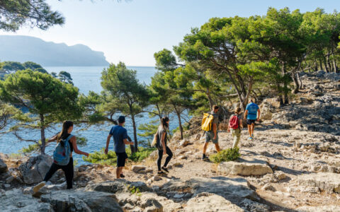 Small group of hikers walks over beige stones by green pine trees beside teal sea on a luxury cruise in the French Riviera.