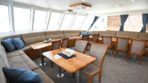 Dining room of Heritage Explorer small ship with wraparound beige bench seats, wooden 4-top tables & chairs & wall of windows.