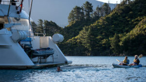 Aft of New Zealand yacht Heritage Explorer with large, water-line swim step & guests kayaking & swimming in calm, sunny sea.