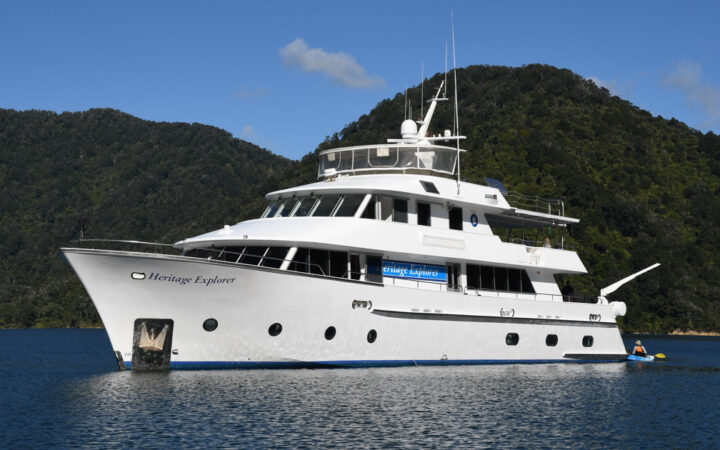 Heritage Explorer white yacht with 4 decks sits in sunny, calm sea backed by dense green hills under a blue sky in New Zealand.