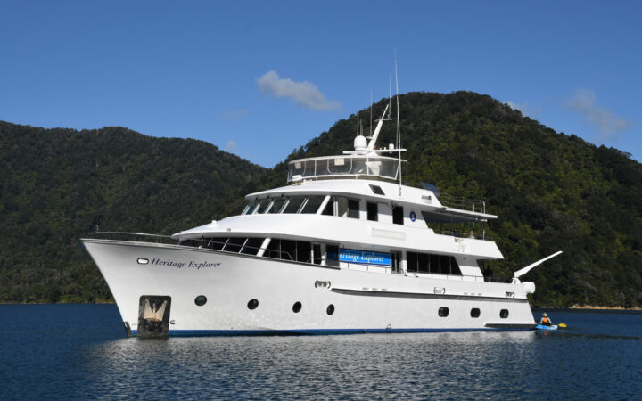 Heritage Explorer white yacht with 4 decks sits in sunny, calm sea backed by dense green hills under a blue sky in New Zealand.