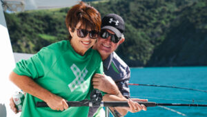 Man & woman fish from the deck of Heritage Explorer yacht on a sunny day with teal sea & green hills in New Zealand.