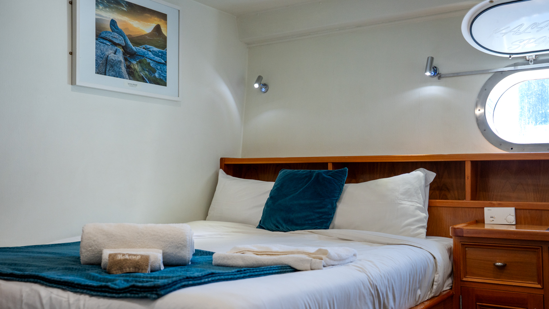 Salvin's cabin with double bed in blue & white with wood headboard & side table under large porthole on Heritage Explorer.