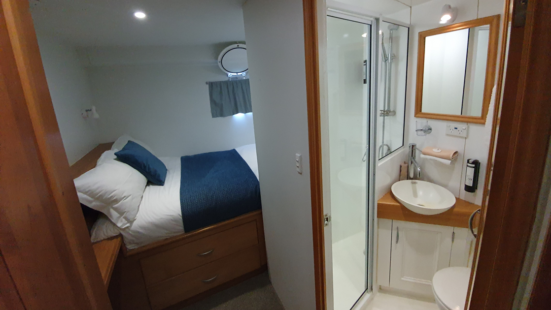 Wandering cabin with twin bed in blue & white with wood headboard & side table, porthole & small bathroom on Heritage Explorer.