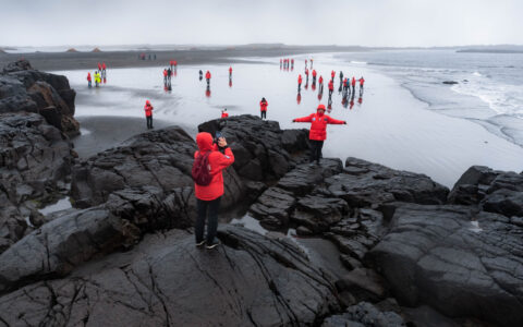 Iceland Circumnavigation travelers in red jackets walk around & pose for photos on a black-sand beach in ocean spray.