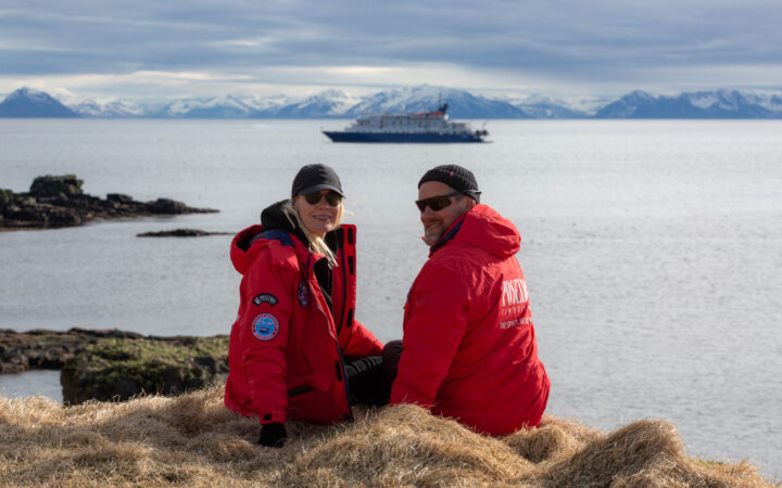 Man & woman in red jackets sit on grassy overlook with small blue & white ship offshore on a sunny day in Iceland.